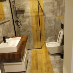 PGbud - Nowoczesna łazienka z kabiną typu walk-in, umywalką nablatową z drewnianym blatem i toaletą podwieszaną. Szare płytki kontrastują z drewnopodobną podłogą.