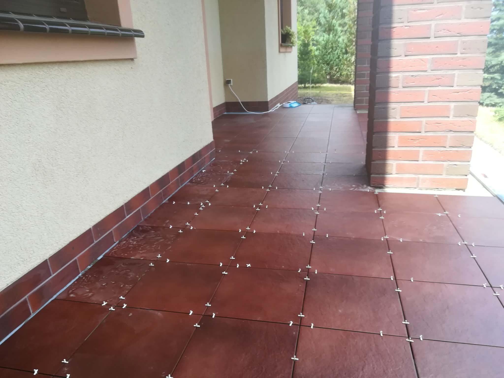 Układanie brązowych płytek na tarasie z użyciem krzyżyków dystansowych. Widoczne fragmenty elewacji i ceglanej kolumny. Prace glazurnicze w toku.