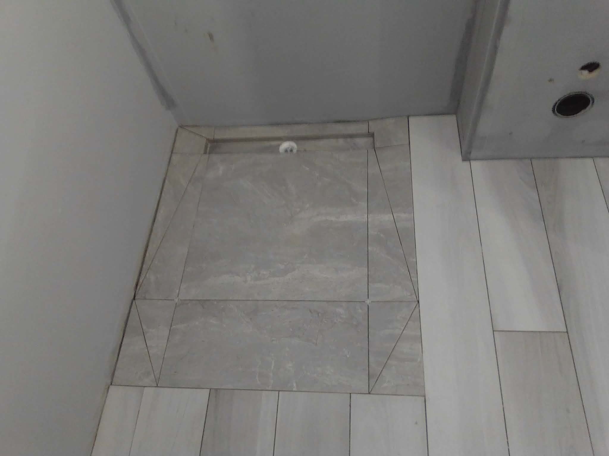 Wykończona strefa prysznicowa z odpływem liniowym i szarymi płytkami imitującymi kamień, ułożonymi w różnych formatach. Widok z góry, surowy stan wykończenia.