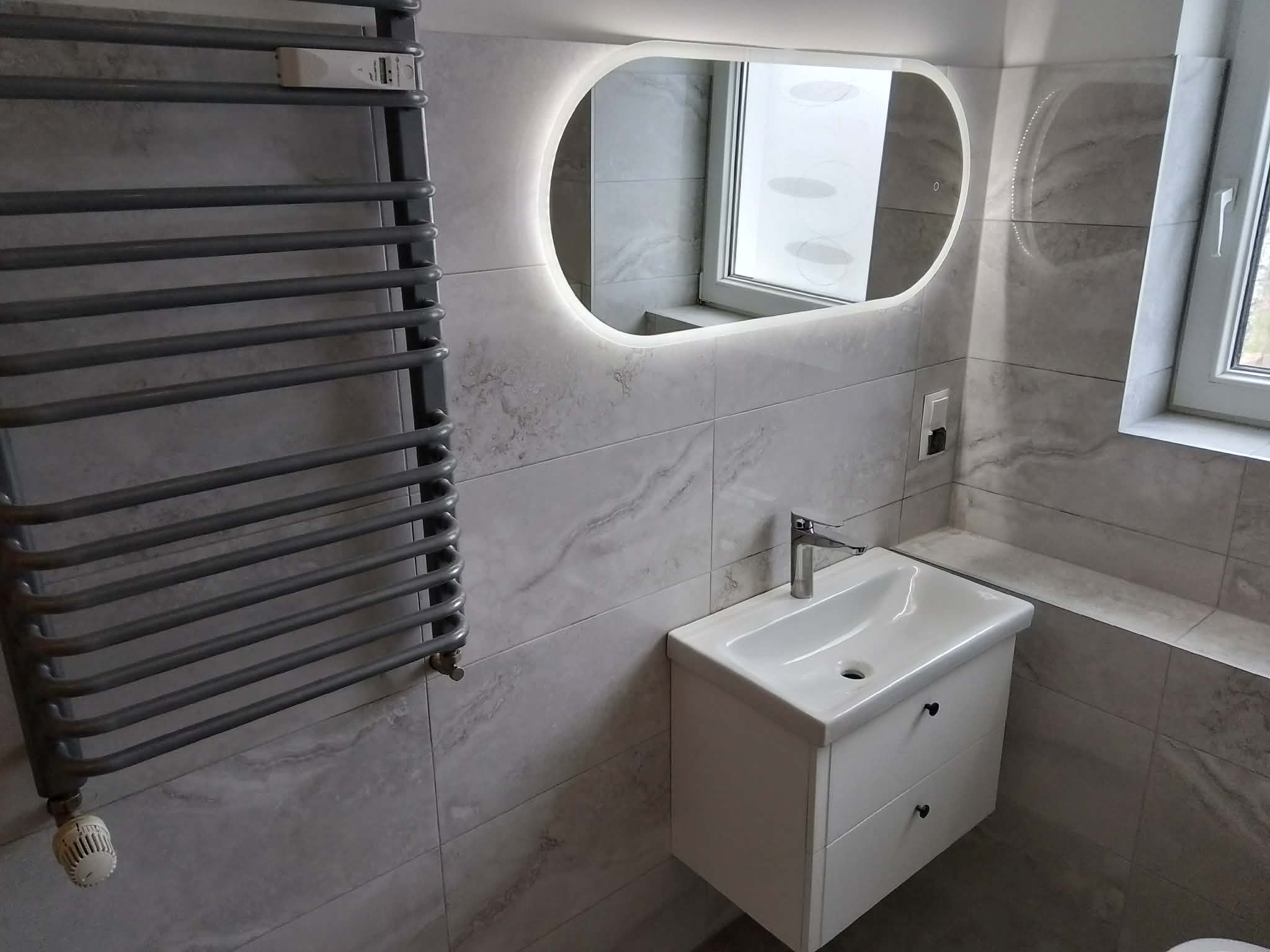 Nowoczesna łazienka z szarymi płytkami imitującymi marmur, podświetlanym lustrem owalnym, grzejnikiem drabinkowym i białą szafką z umywalką.