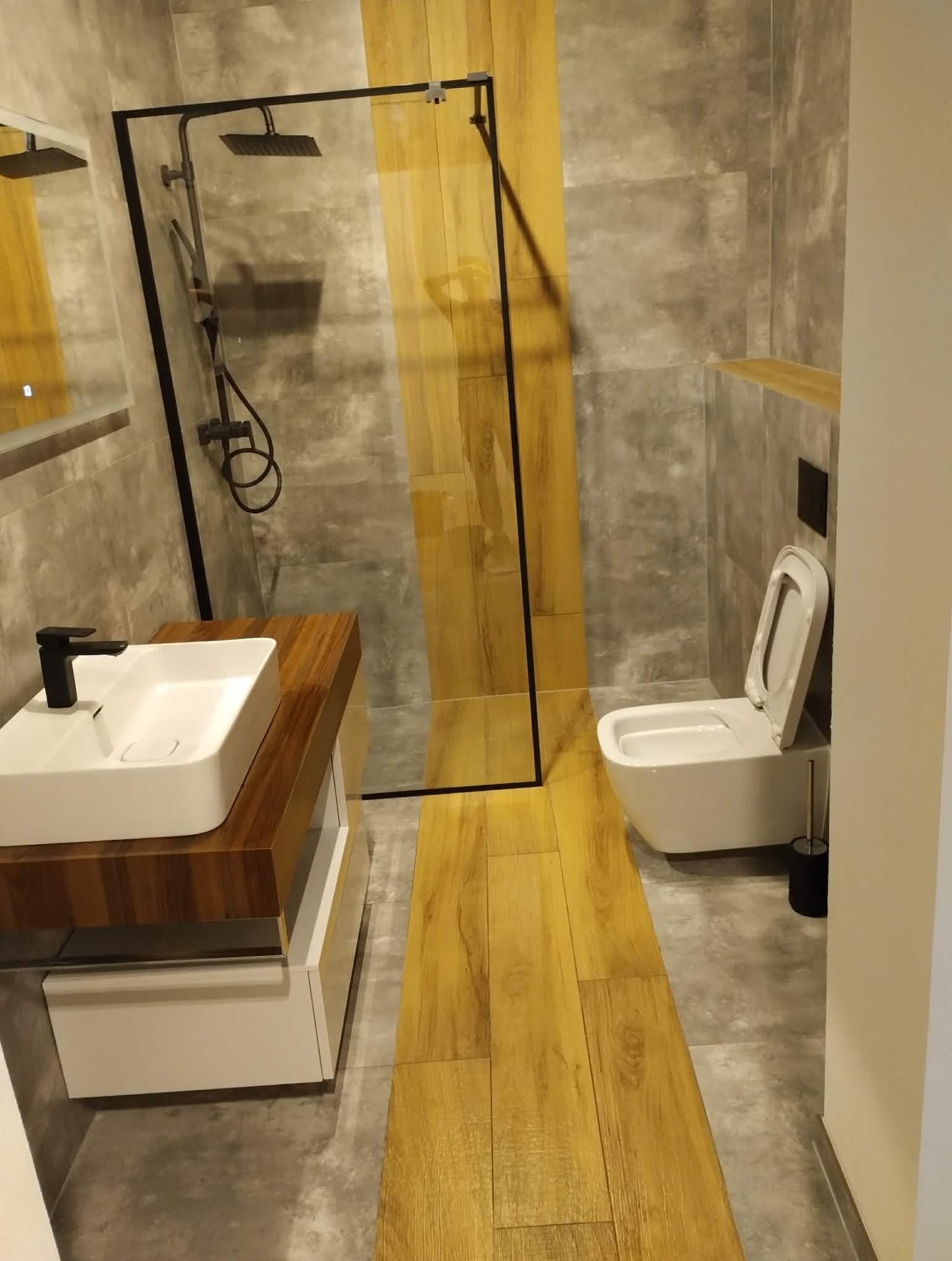 Nowoczesna łazienka z kabiną typu walk-in, umywalką nablatową z drewnianym blatem i toaletą podwieszaną. Szare płytki kontrastują z drewnopodobną podłogą.
