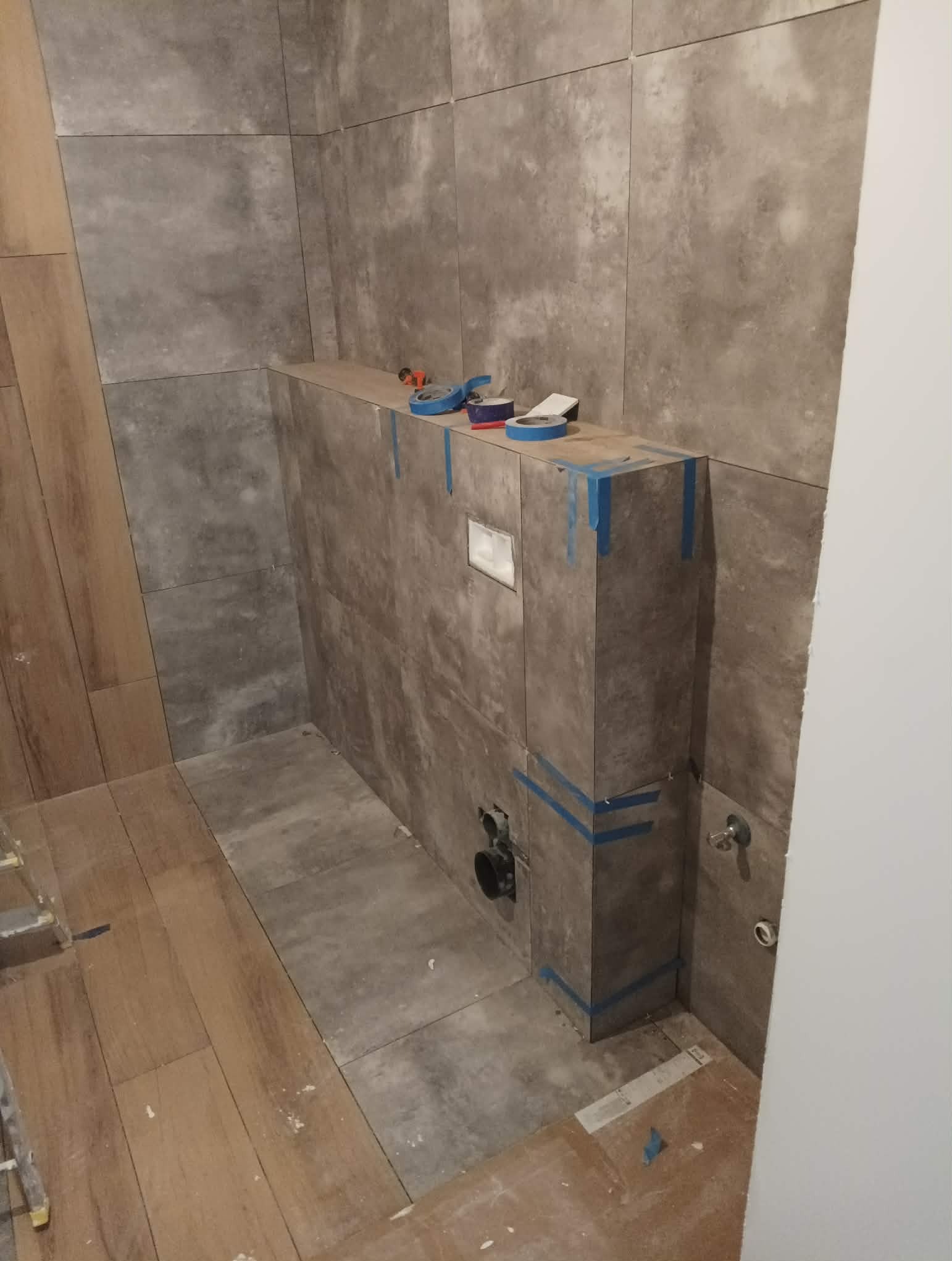 Wnętrze łazienki w trakcie prac glazurniczych. Widoczne płytki imitujące kamień na ścianach i podłodze, obudowa stelaża WC z niebieską taśmą malarską.