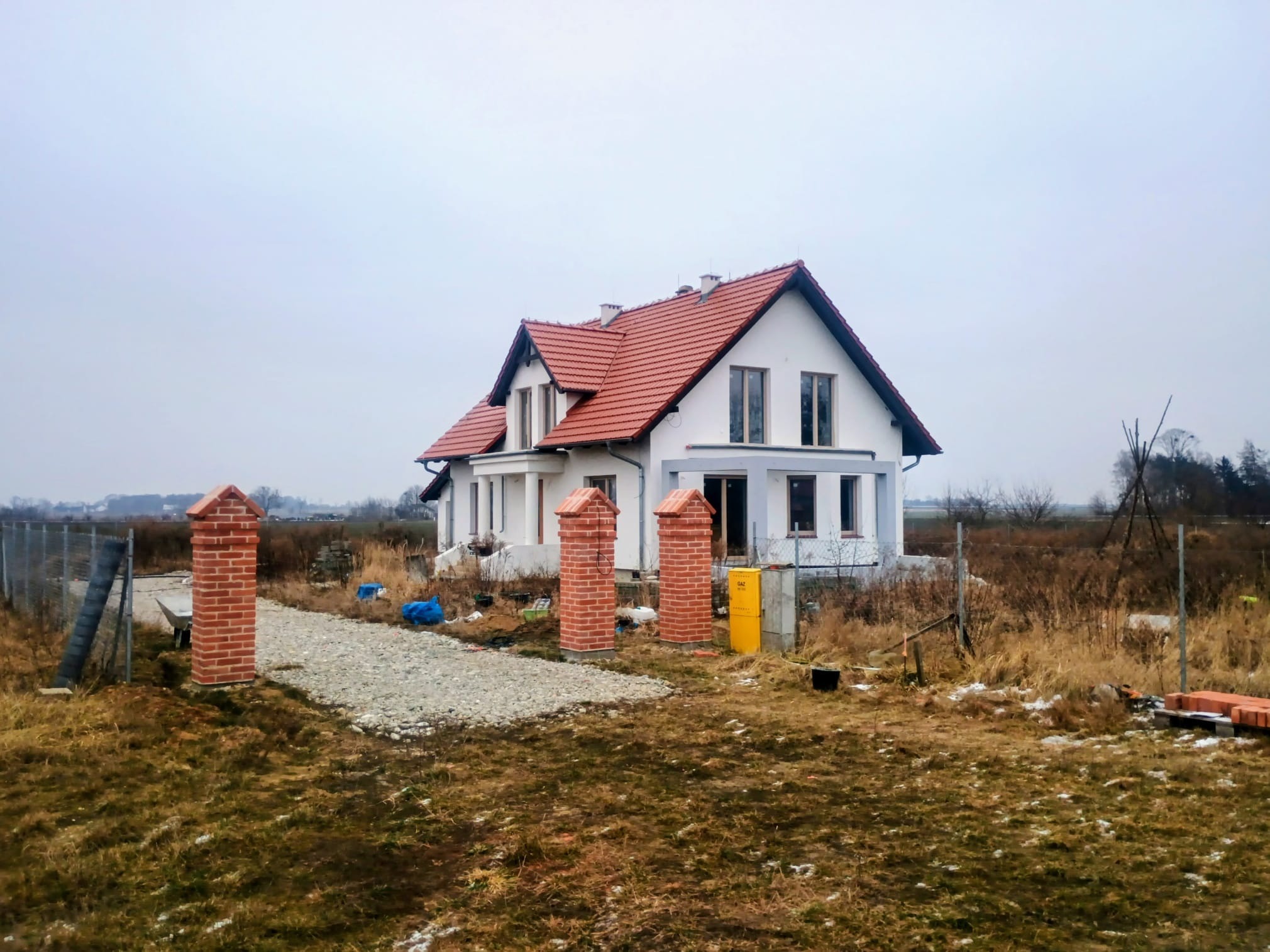 Biały dom z czerwoną dachówką w trakcie budowy, widoczne ceglane słupki wejściowe i teren budowy. Pochmurne niebo w tle.
