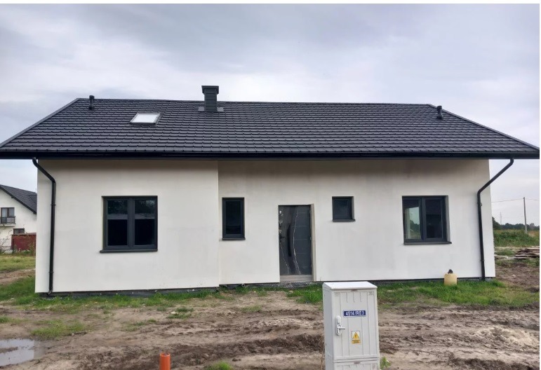 Nowoczesny, parterowy dom z ciemną dachówką i grafitową stolarką okienną. Biała elewacja, widoczny fragment skrzynki elektrycznej. Ujęcie z poziomu gruntu.