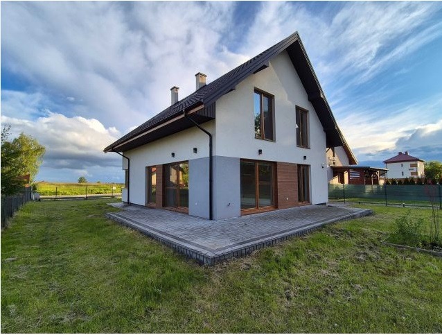 Dwukondygnacyjny dom z dwuspadowym dachem, wykończony w bieli i szarości, z drewnianymi akcentami, otoczony trawnikiem i kostką brukową. Widok z niskiej perspektywy.