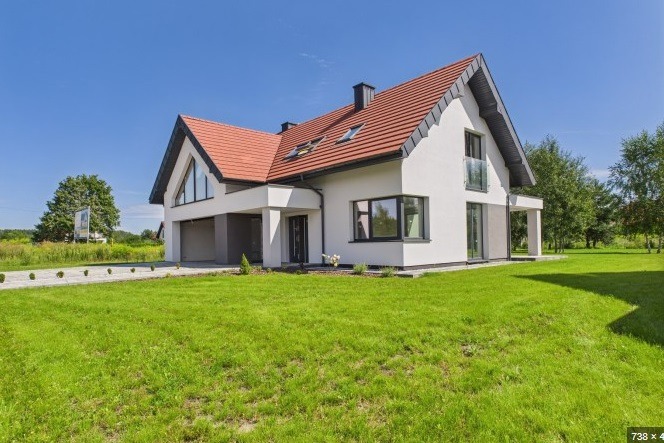 Nowoczesny dom z czerwoną dachówką i białymi ścianami, garaż w bryle budynku, duże okna, zadbany trawnik, błękitne niebo. Ujęcie z poziomu trawnika.