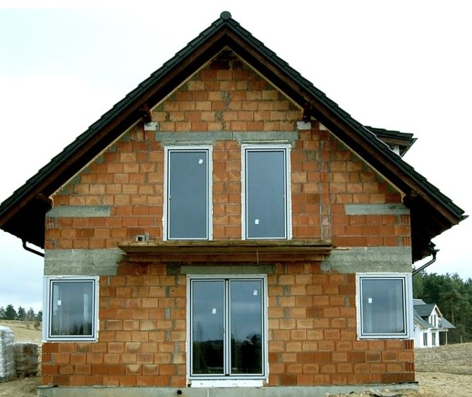 Niedokończony dom z czerwonej cegły z zamontowanymi oknami. Widoczne szare nadproża i czarny dach. W tle drzewa i fragment innego budynku.