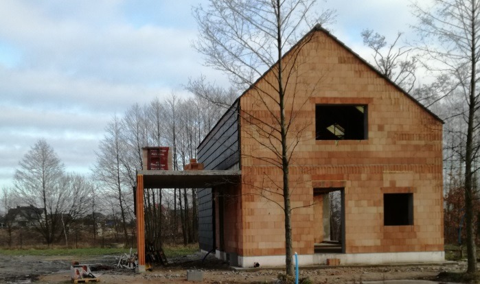 Surowy, dwukondygnacyjny dom z cegły w trakcie budowy, z widocznymi otworami okiennymi i drzwiowymi, na tle drzew i pochmurnego nieba. Częściowo zadaszony.