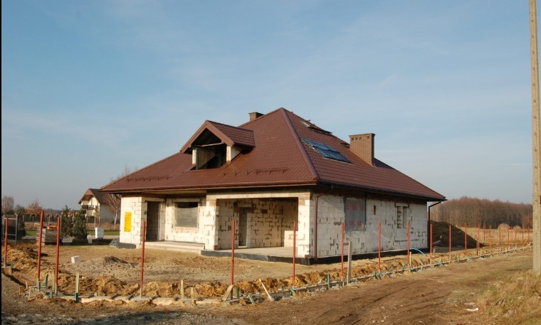 Niedokończony dom jednorodzinny z brązowym dachem, widoczny z zewnątrz. Ściany z białego pustaka, częściowo zakryte folią. W tle błękitne niebo i drzewa.