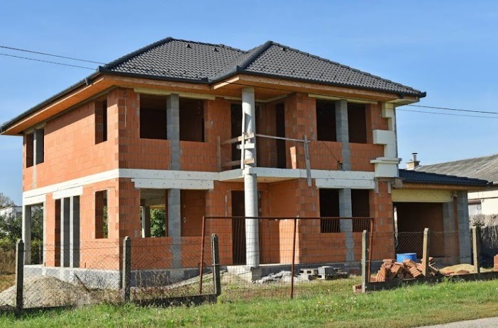 Dwukondygnacyjny dom w trakcie budowy z czerwonej cegły, z widocznymi betonowymi słupami i czarnym dachem, otoczony siatką ogrodzeniową na tle błękitnego nieba.