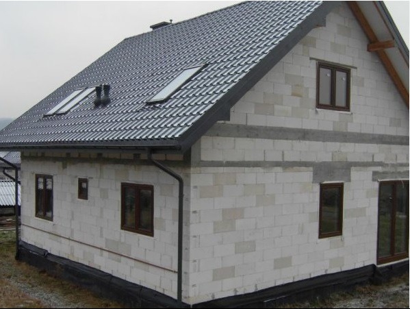 Surowy dom z białego bloczka, z ciemną dachówką i oknami. Widoczne detale: rynny, okna dachowe, izolacja fundamentów. Budynek w trakcie budowy, z widocznymi spoinami.