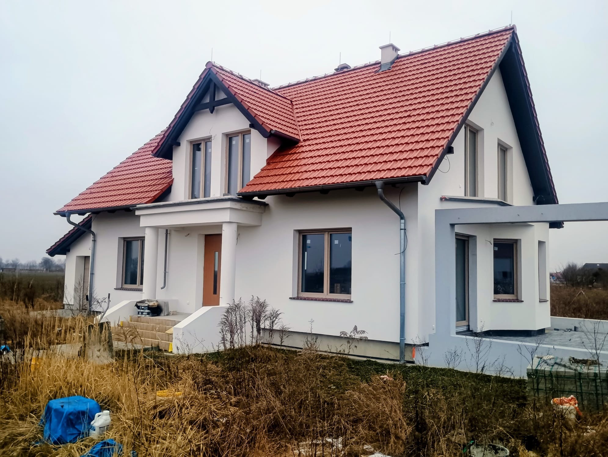 Biały dom jednorodzinny z czerwoną dachówką, widoczny z zewnątrz, z elementami wykończeniowymi i fragmentem terenu przed budynkiem. Ujęcie w pochmurny dzień.