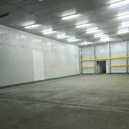NT Technologia - Pusta hala magazynowa z metalowymi ścianami i betonową podłogą, oświetlona lampami jarzeniowymi. Widoczne żółte regały i otwarte drzwi w głębi.