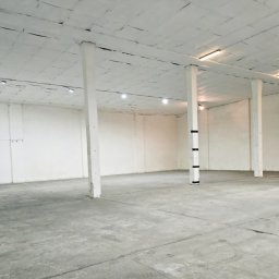 NT Technologia - Puste, jasne wnętrze magazynu z betonową podłogą i białymi ścianami, wsparte na kolumnach. Widoczne oświetlenie sufitowe i ślady użytkowania na powierzchniach.