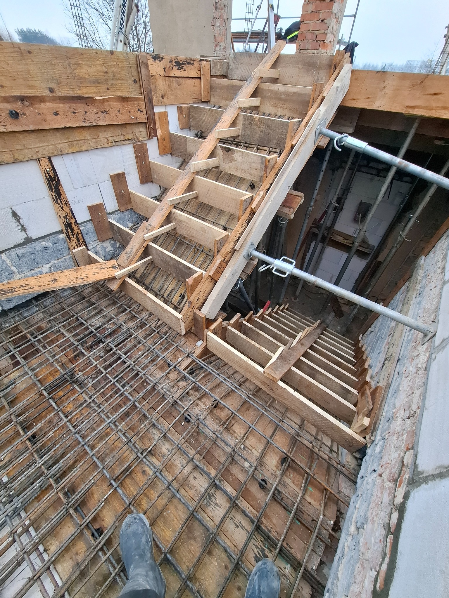 Zbrojenie i szalunek schodów na budowie domu, widok z góry. Metalowa siatka zbrojeniowa na drewnianym deskowaniu, drewniane formy schodów, fragment muru z cegły.
