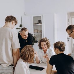 Kursy Osteopatyczne - Grupa osób w jasnym pomieszczeniu, uczestnicząca w spotkaniu lub szkoleniu. Kobieta z kręconymi włosami gestykuluje, uśmiechając się do rozmówców.