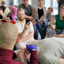 Kursy Osteopatyczne - Kurs masażu: terapeuta w rękawiczkach uciska punkty na twarzy leżącego pacjenta, obserwatorzy z telefonami w tle. Okulary na głowie terapeuty.