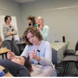 Kursy Osteopatyczne - Kurs masażu: instruktorka demonstruje technikę na leżącej modelce, grupa obserwuje. Widoczne igły do akupunktury, stół do masażu i osoby w tle.