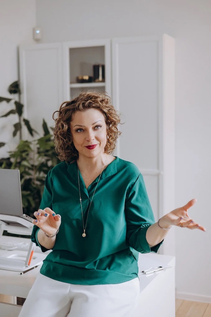 Kobieta w szmaragdowej bluzce gestykuluje, siedząc przy biurku z laptopem. Jasne wnętrze, naturalne światło, profesjonalny wizerunek trenera/szkoleniowca.
