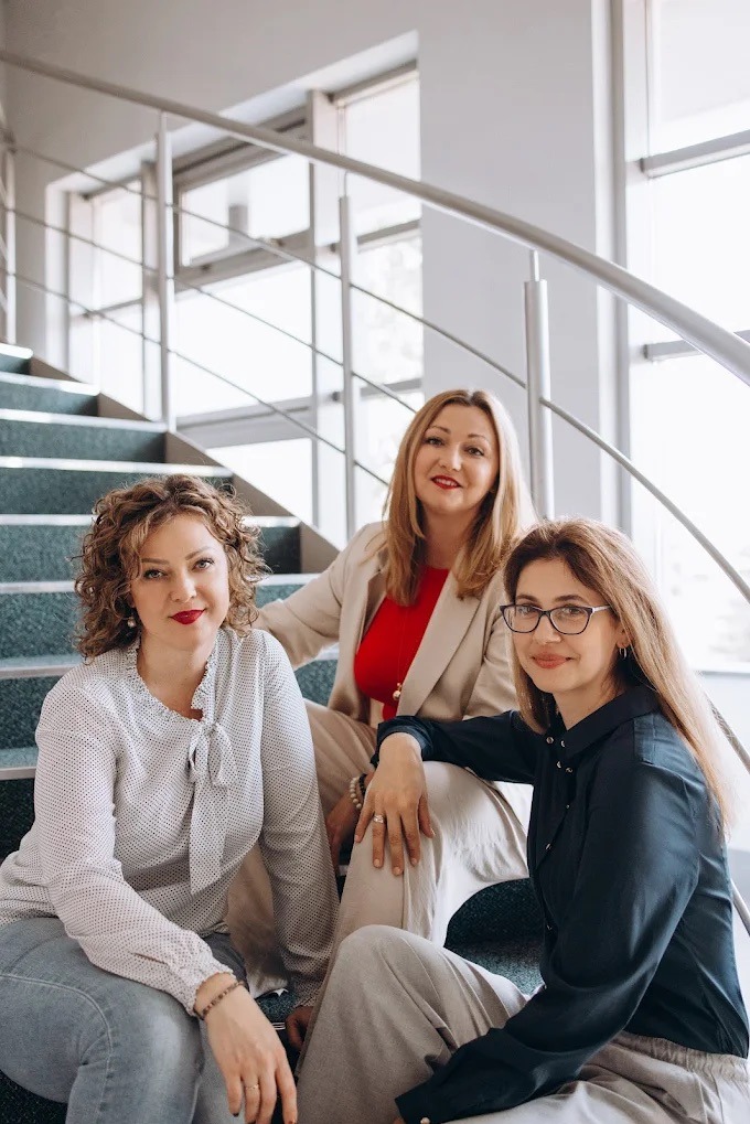 Trzy uśmiechnięte kobiety siedzą na schodach w jasnym wnętrzu. Kobiety ubrane w eleganckie bluzki i spodnie, sugerujące profesjonalne środowisko szkoleniowe.