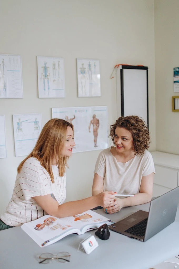 Dwie kobiety przy biurku z laptopem i otwartą książką z ilustracjami anatomicznymi. W tle plakaty z układem kostnym. Jasne wnętrze, możliwe szkolenie medyczne.