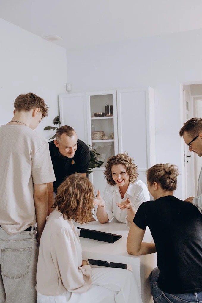 Grupa osób w jasnym pomieszczeniu, uczestnicząca w spotkaniu lub szkoleniu. Kobieta z kręconymi włosami gestykuluje, uśmiechając się do rozmówców.