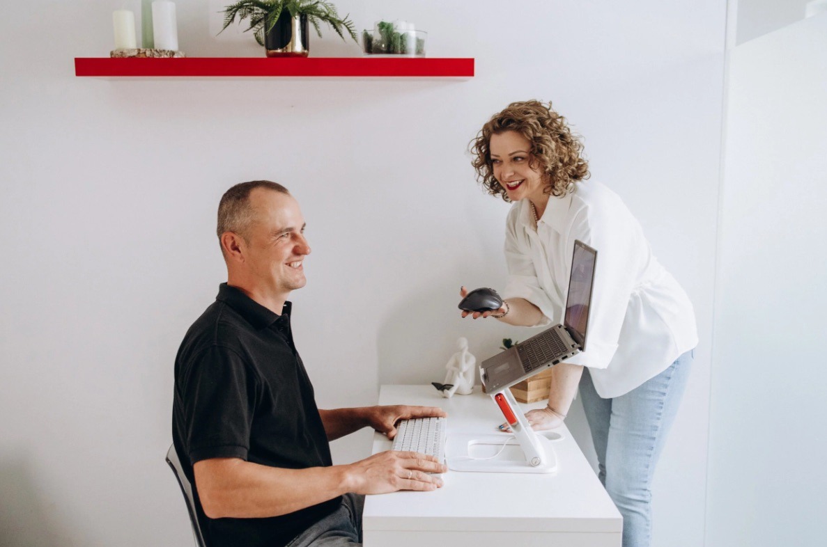 Uśmiechnięta kobieta z myszką w dłoni instruuje mężczyznę siedzącego przy biurku z laptopem na białym, regulowanym stojaku. Czerwona półka z dekoracjami w tle.