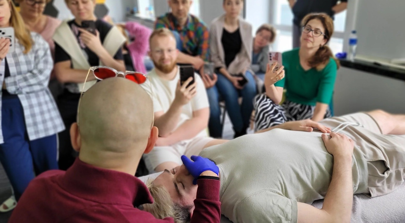 Kurs masażu: terapeuta w rękawiczkach uciska punkty na twarzy leżącego pacjenta, obserwatorzy z telefonami w tle. Okulary na głowie terapeuty.