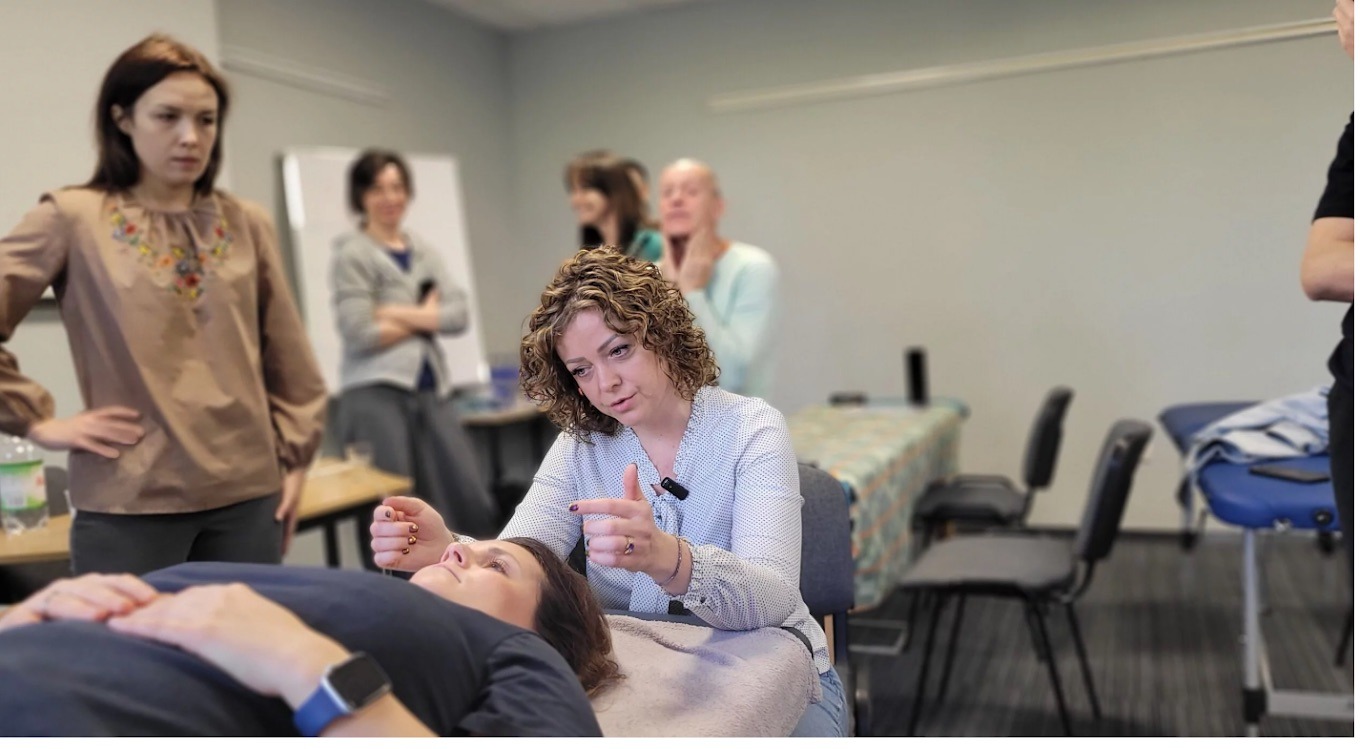Kurs masażu: instruktorka demonstruje technikę na leżącej modelce, grupa obserwuje. Widoczne igły do akupunktury, stół do masażu i osoby w tle.
