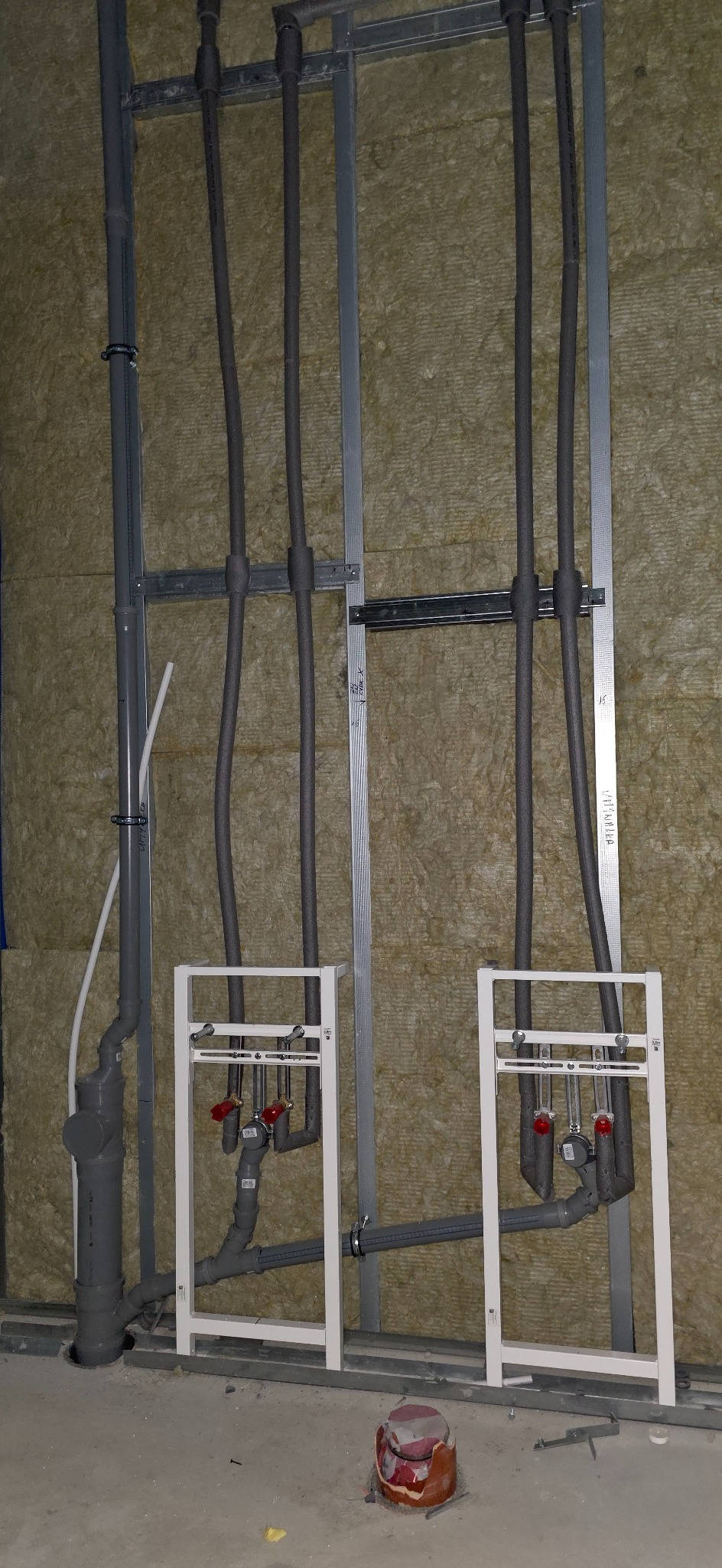 Szara instalacja wodno-kanalizacyjna w stelażu, rury w izolacji, widoczne zawory z czerwonymi pokrętłami, ściana z izolacją z wełny mineralnej.