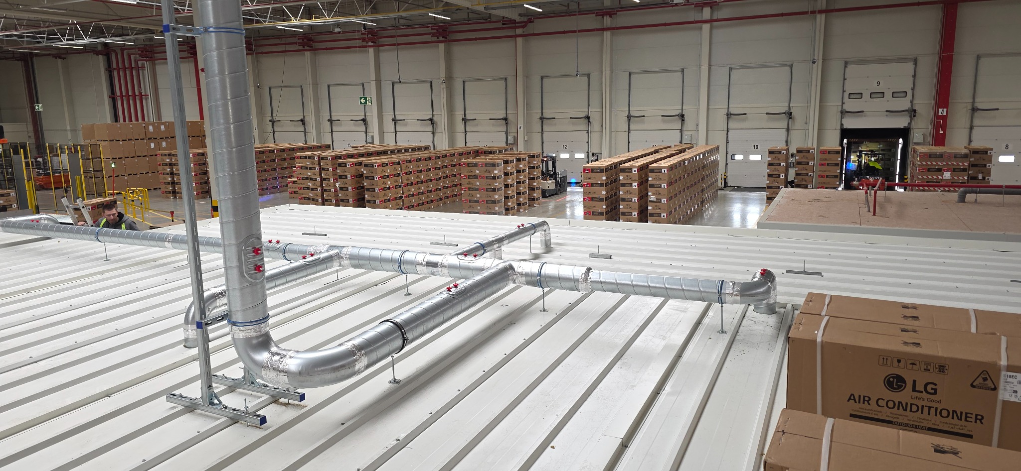 Instalacja wentylacyjna z rurami na dachu hali produkcyjnej, widoczne kartony z klimatyzatorami LG i pracownik na drabinie. Metalowe rury wentylacyjne na białym dachu.