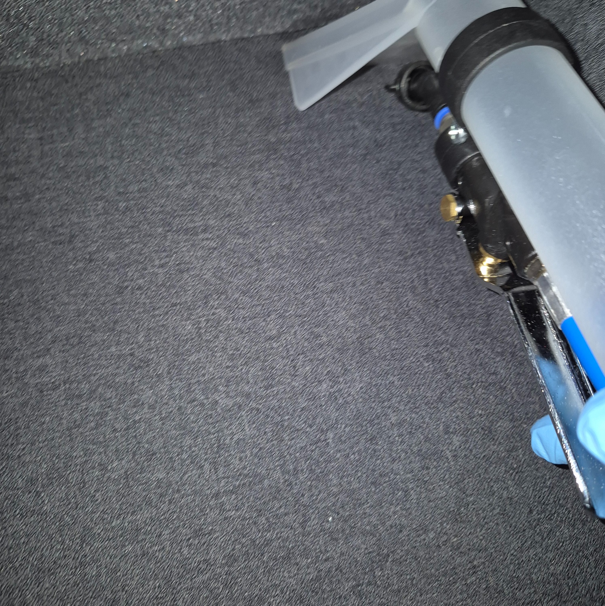 Czyszczenie ciemnej tapicerki tekstylnej za pomocą profesjonalnego ekstraktora. Widoczna dysza i fragment dłoni w niebieskiej rękawicy ochronnej.