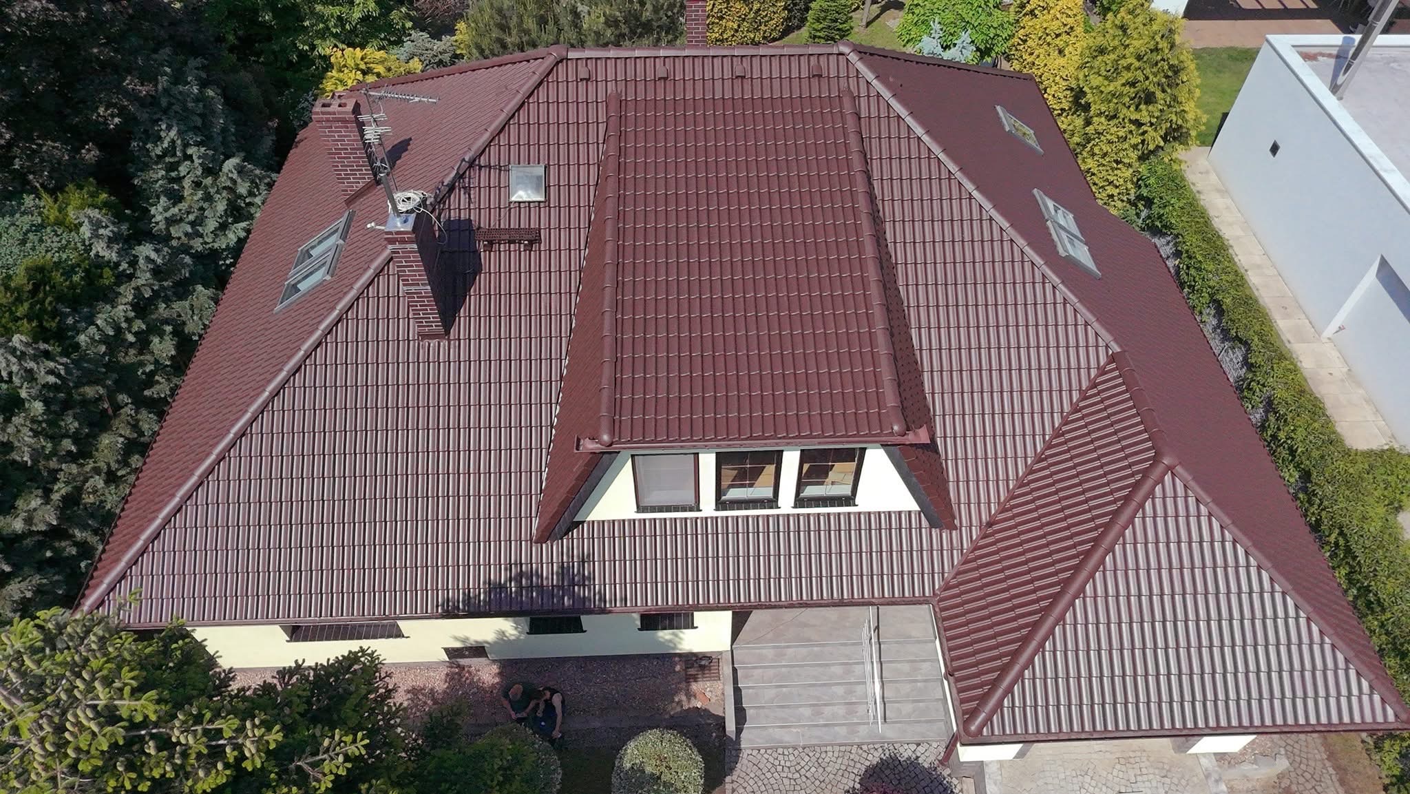 Widok z góry na duży dom z brązowym dachem z dachówki ceramicznej, kominem i oknami dachowymi. Widoczne drzewa i fragment elewacji w odcieniach beżu.