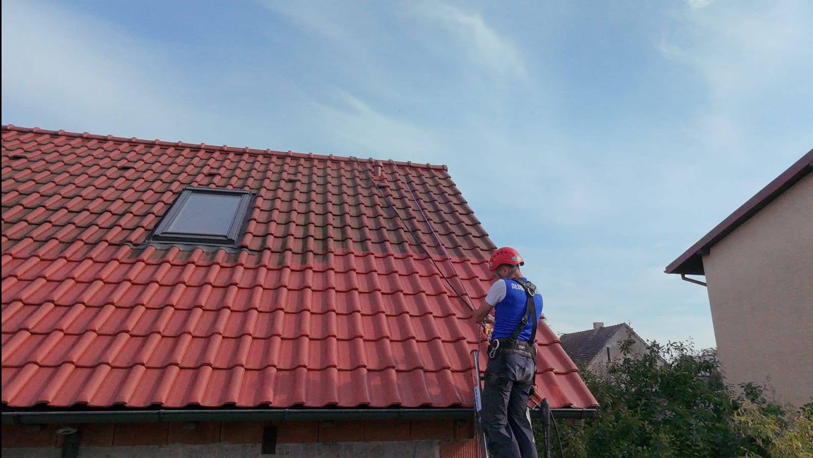 Pracownik w kasku, na drabinie, czyści czerwoną dachówkę z okienkiem. Część dachu jest już umyta, widoczna różnica w kolorze. Błękitne niebo w tle.