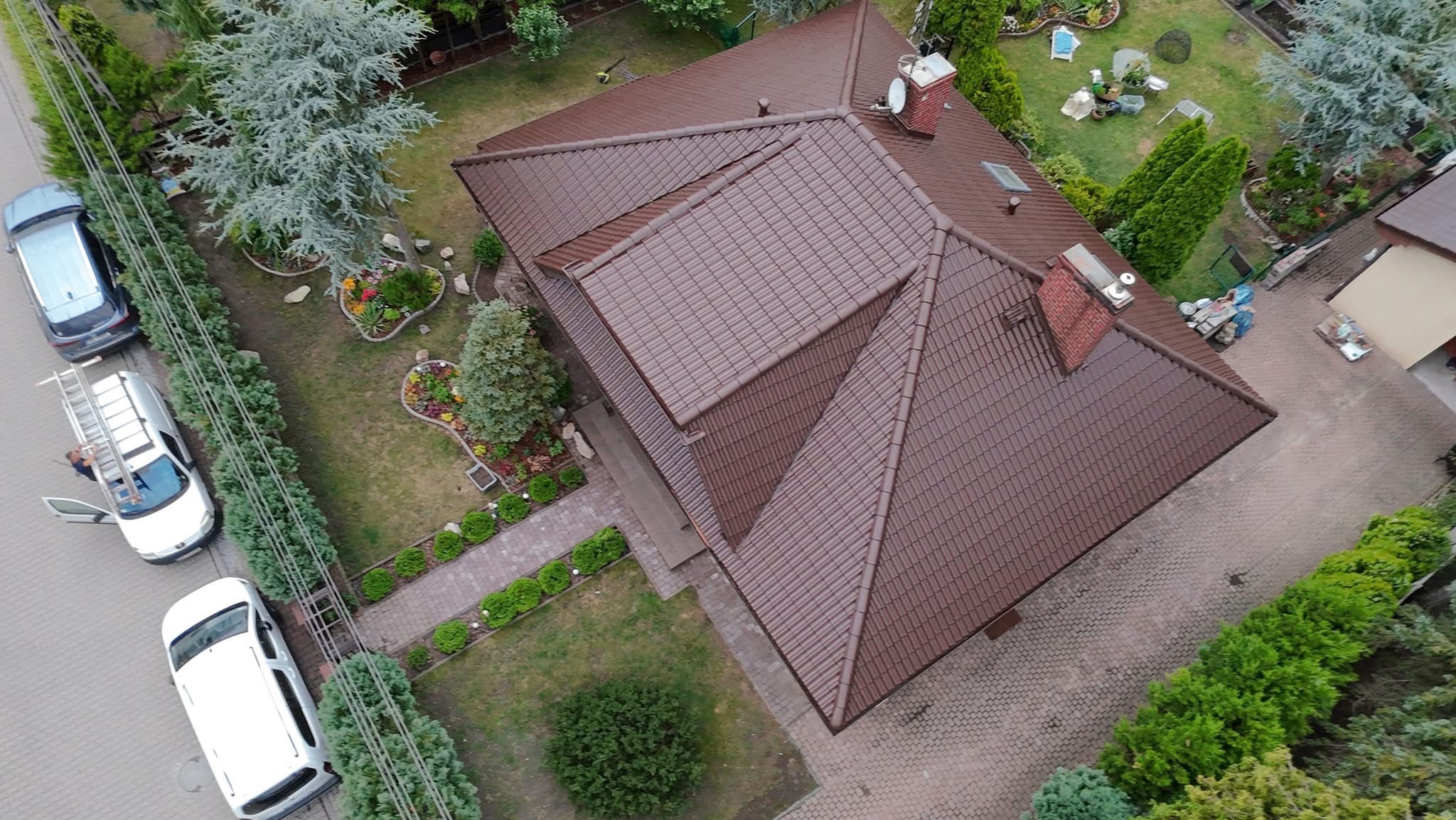 Widok z góry na dom z brązowym dachem, zadbanym ogrodem i podjazdem z kostki. Na ulicy zaparkowane samochody, w tym jeden z rozłożoną drabiną.