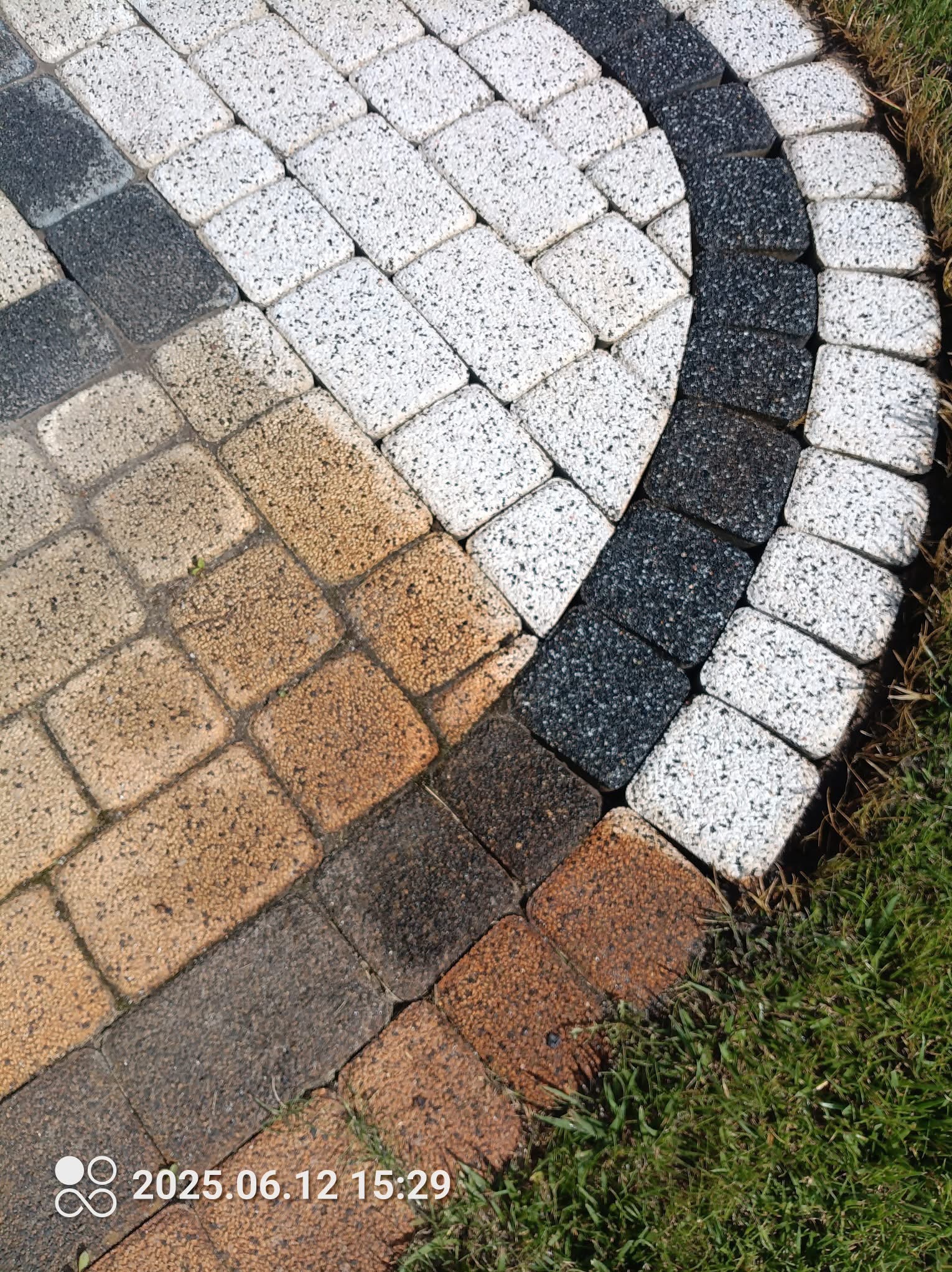 Kolorowa kostka brukowa ułożona w łukowaty wzór, otoczona trawą. Widoczne odcienie szarości, bieli i brązu. Detal nawierzchni z bliska.