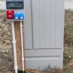 mPower Mieszko Bilski - Szara skrzynka elektryczna z gniazdami i licznikiem, zamontowana na drewnianej podstawie, z widocznymi przewodami w ziemi. Ujęcie z bliska, na zewnątrz.