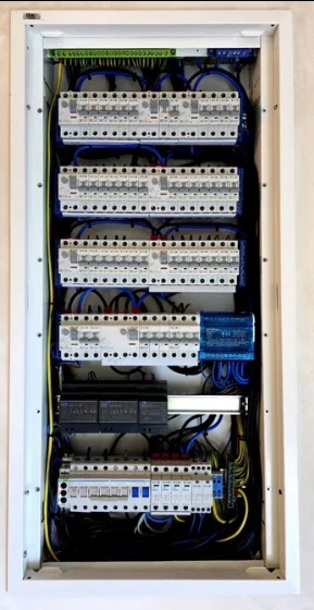 Rozdzielnica elektryczna w białej obudowie, z widocznymi bezpiecznikami, przewodami i innymi elementami instalacji. Układ schludny, przewody uporządkowane.