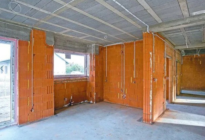 Surowy stan wnętrza z ceglanymi ścianami i widocznymi przewodami elektrycznymi, okno z widokiem na zieleń, betonowa podłoga, sufit z betonowych płyt.