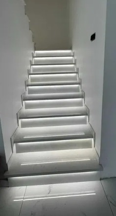 Nowoczesne schody z podświetleniem LED na każdym stopniu, białe ściany i podłoga z połyskiem, czarny włącznik na ścianie, widok z dołu.