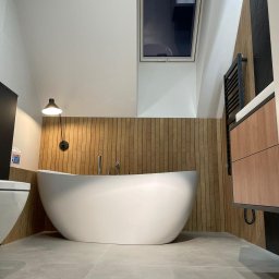 PHU ŻU-RAF RAFAŁ ŻULIŃSKI - Nowoczesna łazienka na poddaszu z wolnostojącą wanną, drewnianą boazerią i oknem dachowym. Minimalistyczny design, szare płytki podłogowe, czarny grzejnik drabinkowy.