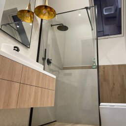 PHU ŻU-RAF RAFAŁ ŻULIŃSKI - Nowoczesna łazienka z kabiną prysznicową typu walk-in, drewnianą szafką pod umywalką i złotymi lampami wiszącymi. Widoczne okno i toaleta.