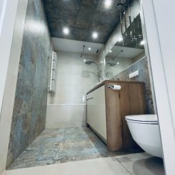 PHU ŻU-RAF RAFAŁ ŻULIŃSKI - Nowoczesna łazienka z płytkami imitującymi kamień na ścianach i podłodze, białą armaturą i drewnianą szafką pod umywalką. Oświetlenie punktowe na suficie.