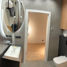 PHU ŻU-RAF RAFAŁ ŻULIŃSKI - Nowoczesna łazienka po remoncie: geometryczne płytki, owalne lustro z podświetleniem LED, drewniane akcenty i wisząca toaleta. Widok na otwarte drzwi do innego pomieszczenia.