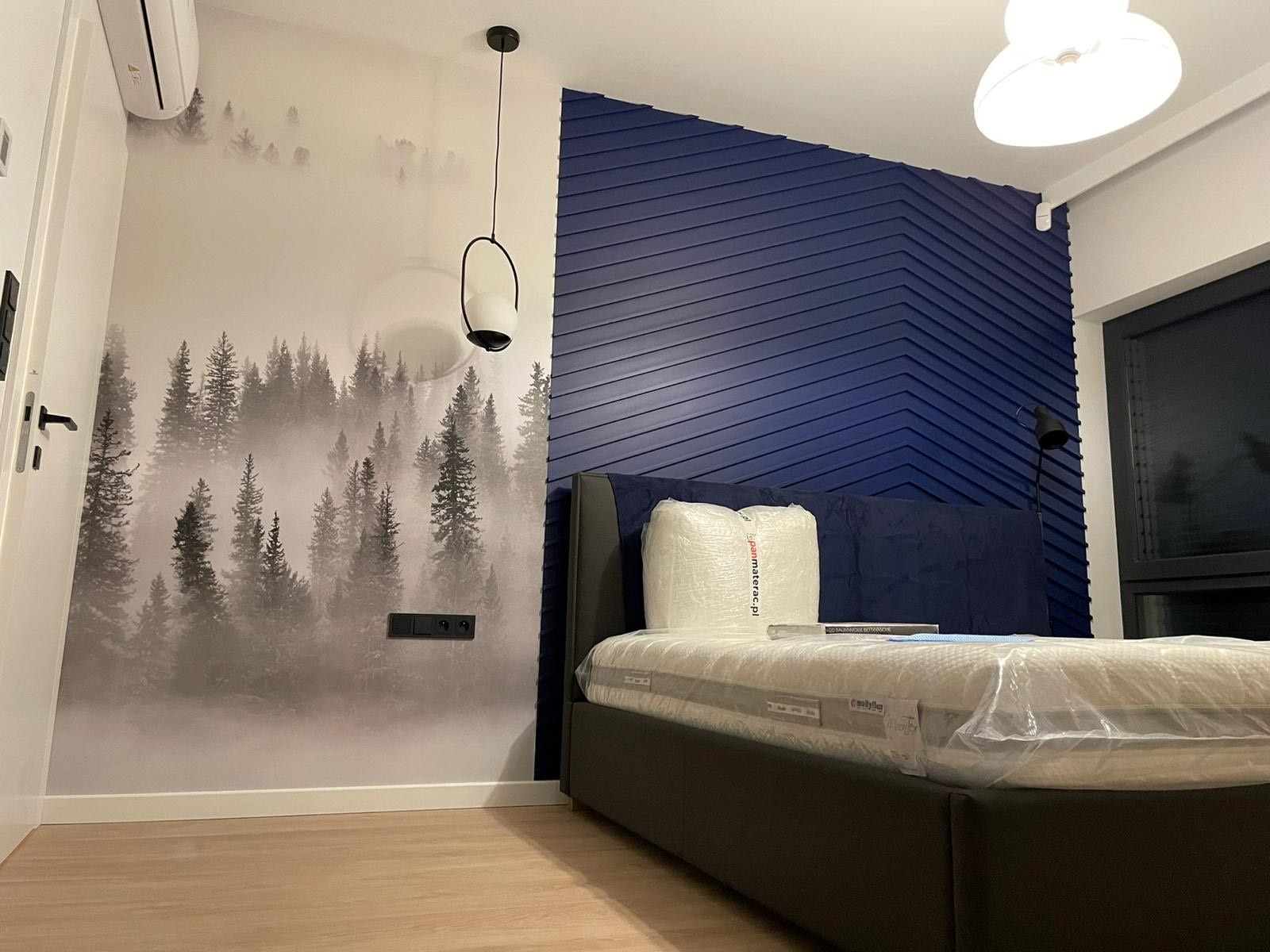 Nowoczesna sypialnia z łóżkiem, fototapetą z motywem lasu i dekoracyjną ścianą w odcieniach granatu. Minimalistyczna lampa wisząca i drewniana podłoga.