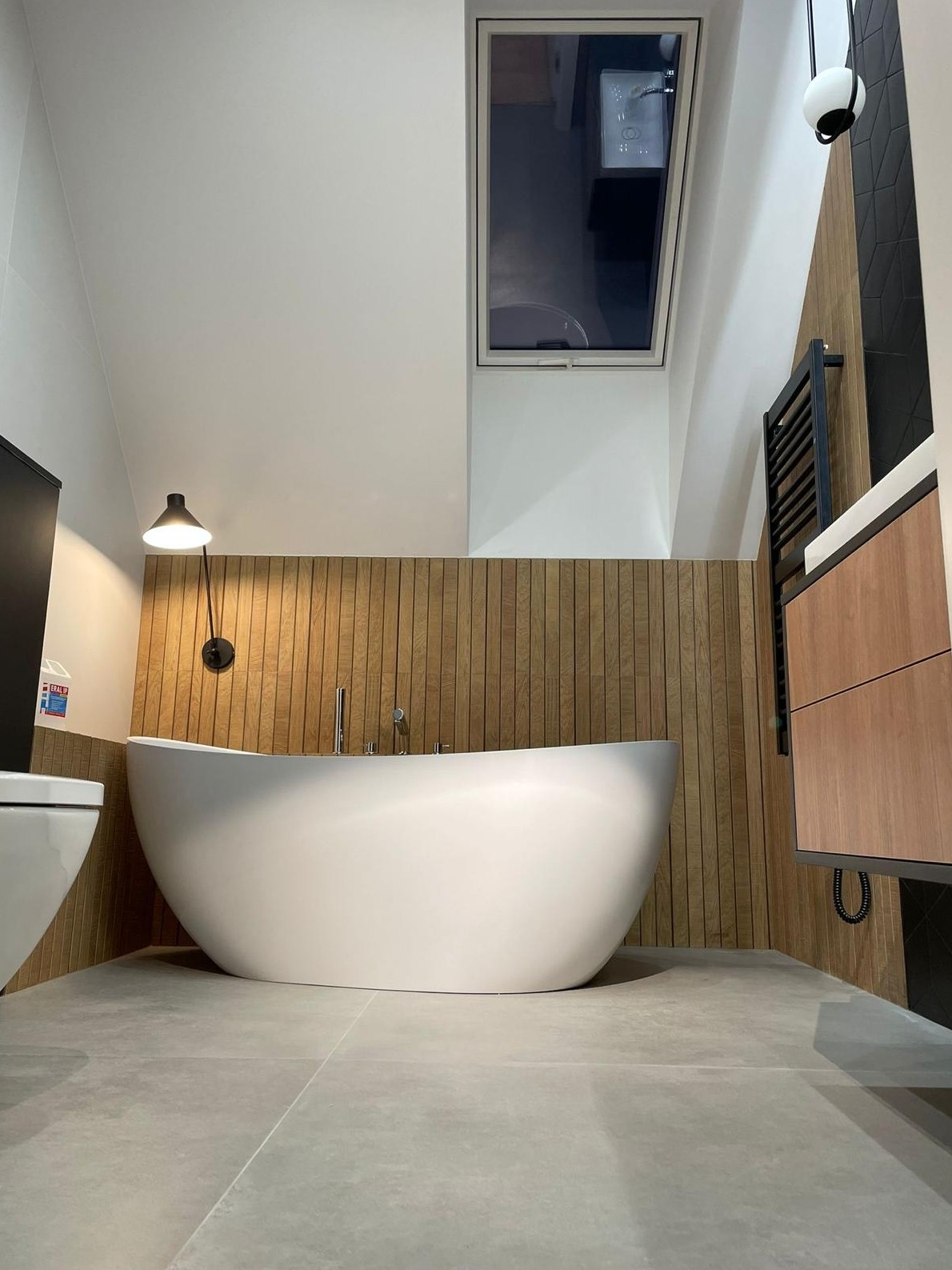 Nowoczesna łazienka na poddaszu z wolnostojącą wanną, drewnianą boazerią i oknem dachowym. Minimalistyczny design, szare płytki podłogowe, czarny grzejnik drabinkowy.