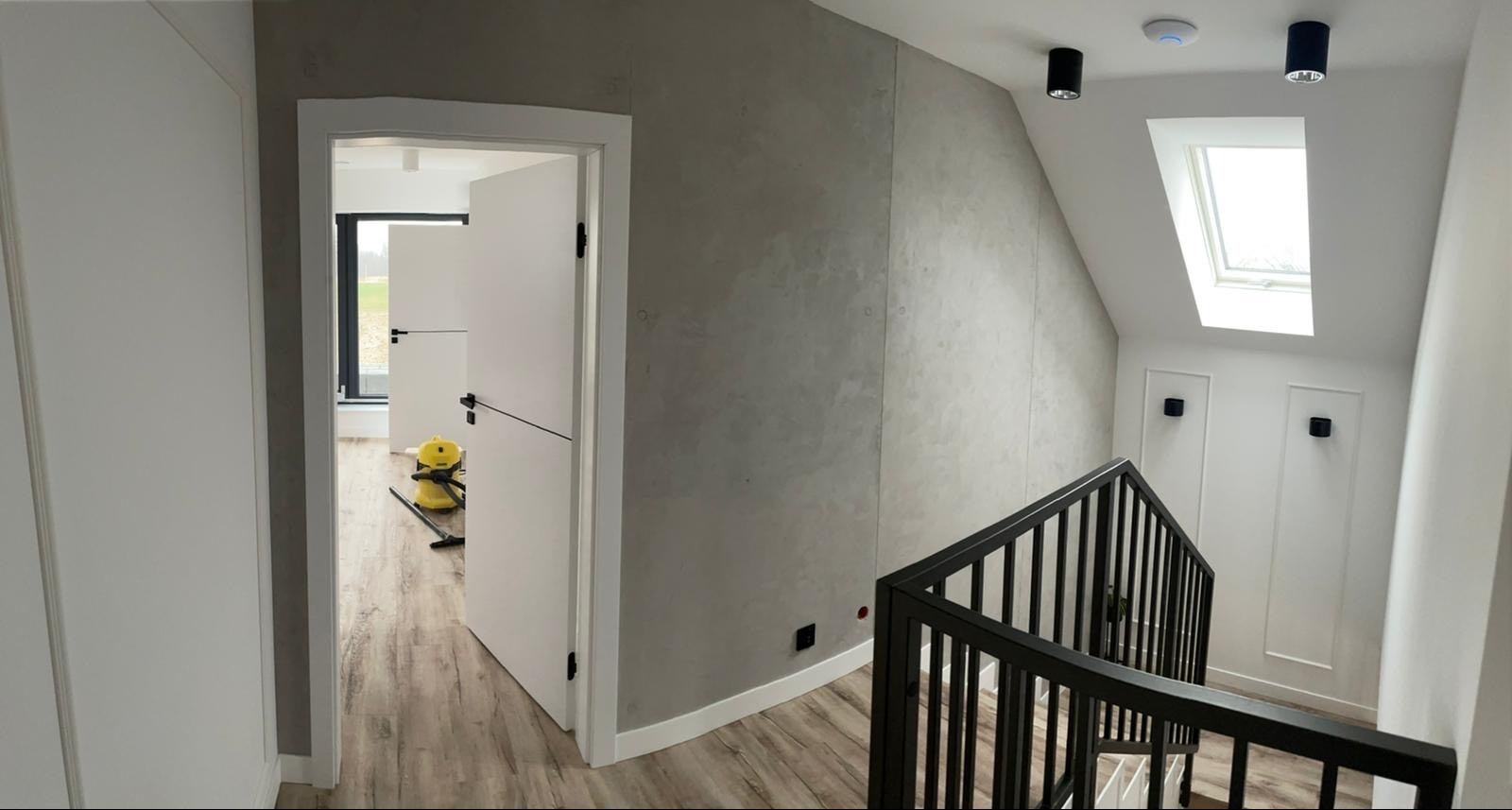 Wykończone wnętrze domu: szara ściana z efektem betonu, białe drzwi z czarnymi klamkami, drewnopodobne panele, czarna balustrada schodów i okno dachowe.