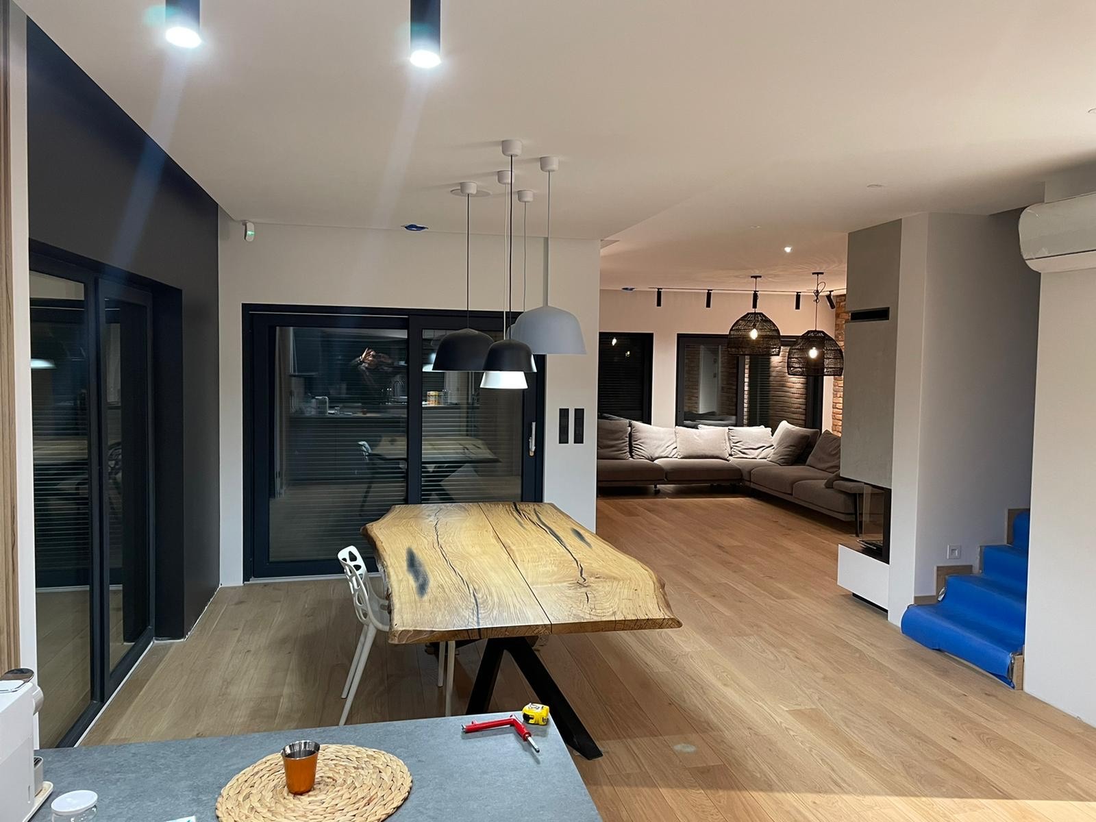 Nowoczesne wnętrze domu z drewnianym stołem o nieregularnym kształcie, parkietem i widokiem na salon z szarą sofą. Schody zabezpieczone folią.
