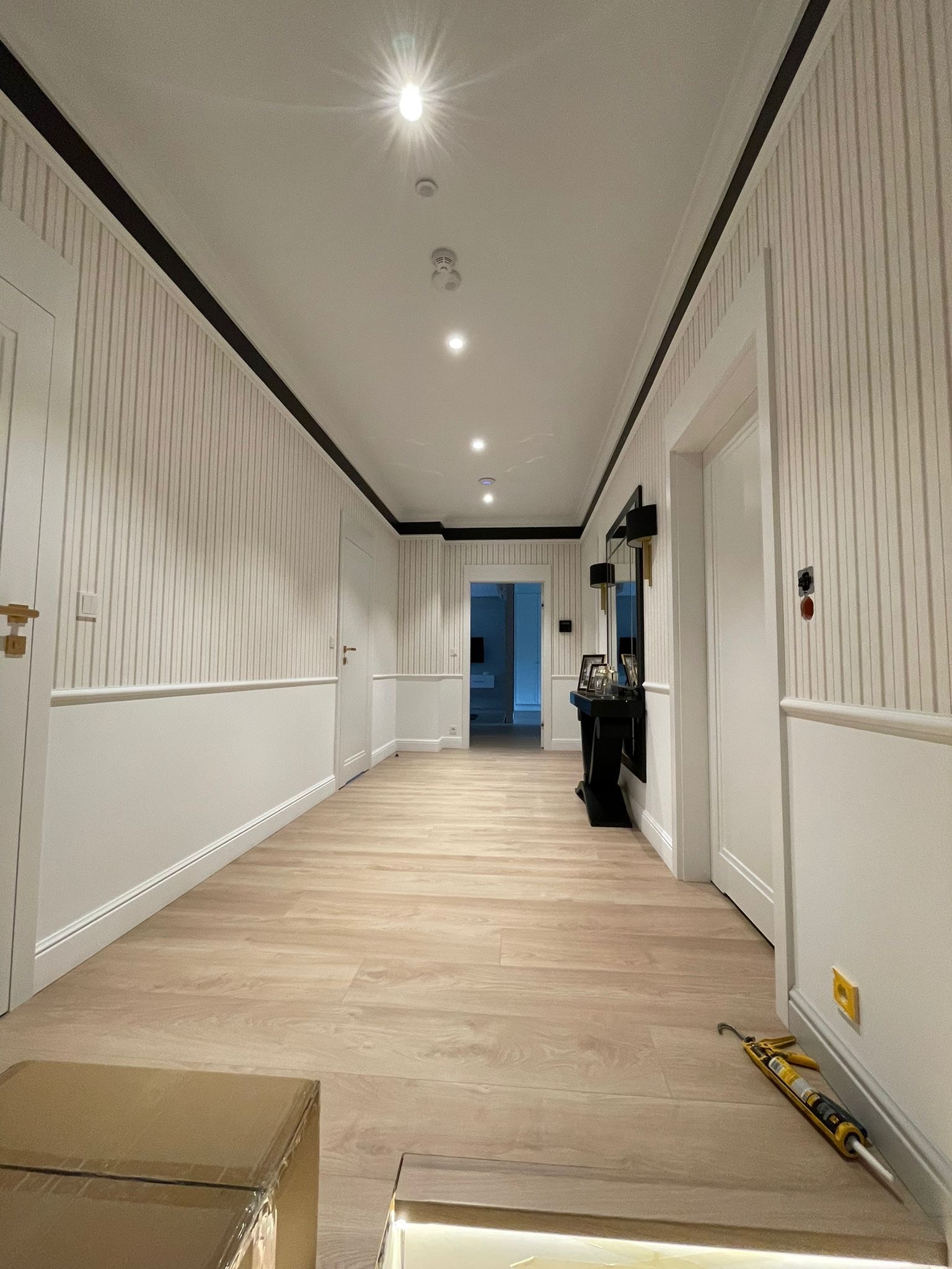 Wykończony korytarz z panelami podłogowymi, tapetą w paski i czarną listwą pod sufitem. Widoczny aplikator do silikonu przy ścianie i karton na podłodze.