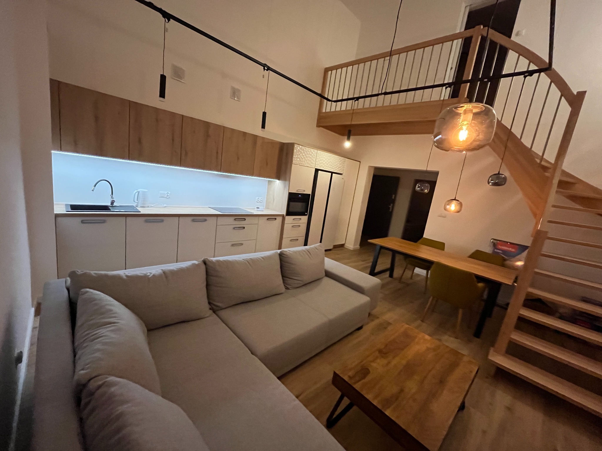 Jasne wnętrze mieszkania z antresolą i drewnianymi schodami. Widoczna kuchnia z oświetleniem LED, stół z krzesłami i narożna sofa. Nowoczesny design z elementami drewna.